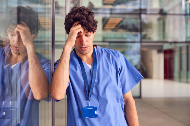 Tired Young Male Doctor Wearing Scrubs Leaning Against Wall In Modern Hospital Building - Foto, imagen