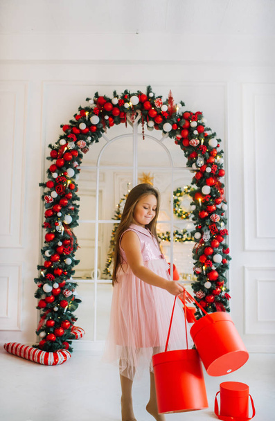 Chica sonriente en vestido festivo de pie cerca del arco de Navidad con cajas de regalo rojas
 - Foto, Imagen