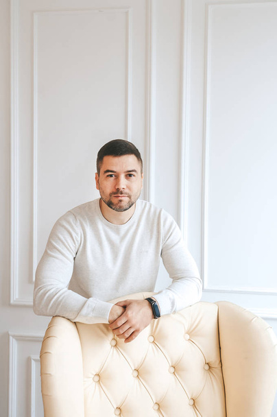 Positive successful young man standing near armchair, dressed white sweater.  - Фото, зображення