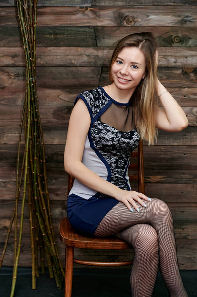 Portrait of a pretty smiling girl with long hair in a gray dress on a background of interior in a room made of wood. Loft style concept. Sits on a chair, shows cute happiness. - Φωτογραφία, εικόνα