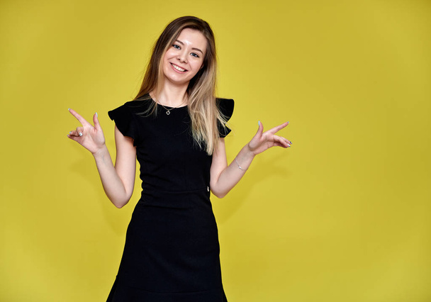 Portrait of a pretty young smiling woman on a yellow background in a black dress with long straight hair. Standing right in front of the camera, Shows emotions, talks in different poses. - Foto, Imagen