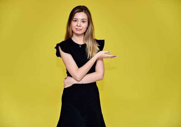 Portrait of a pretty young smiling woman on a yellow background in a black dress with long straight hair. Standing right in front of the camera, Shows emotions, talks in different poses. - Фото, зображення