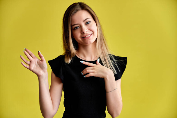 Close-up portrait of a pretty young smiling woman on a yellow background in a black dress with long straight hair. Standing right in front of the camera, Shows emotions, talks in different poses. - Fotografie, Obrázek