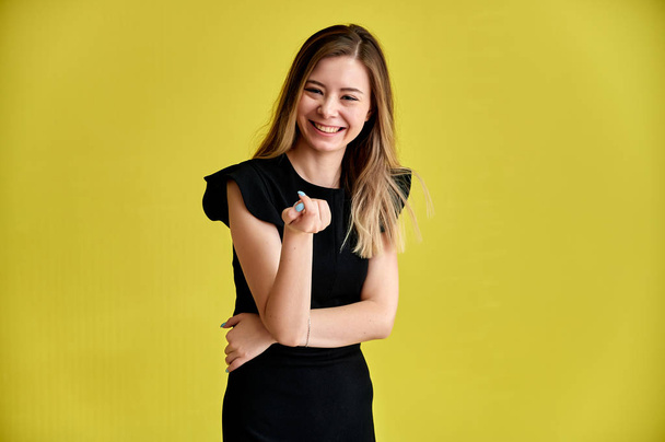 Portrait of a pretty young smiling girl on a yellow background in a black dress with long straight hair. Standing right in front of the camera, Shows emotions, talks in different poses. - Фото, зображення