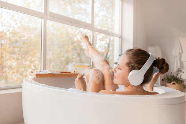 schöne junge Frau hört Musik und trinkt Cocktail, während sie zu Hause badet - Foto, Bild
