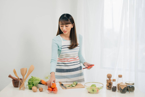 Young woman with vegetables smiling while using mobile phone in kitchen - Photo, Image
