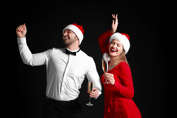 Happy couple in Santa Claus hats and with glasses of champagne on dark background - Photo, Image
