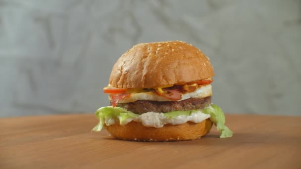 Hampurilainen pyörii puulaudalla. Burger kanssa cutlet salaatti juustoa ja tomaatteja pyörii vastaan harmaa seinä
. - Materiaali, video