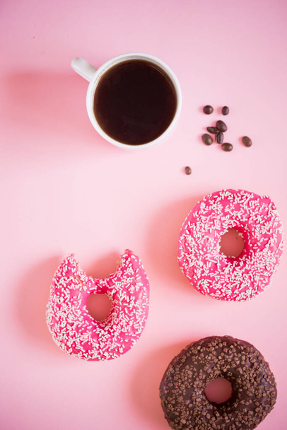 Ízletes és finom fánk rózsaszín cukormázzal és porral, egy csésze aromás kávéval fehér fa alapon. - Fotó, kép