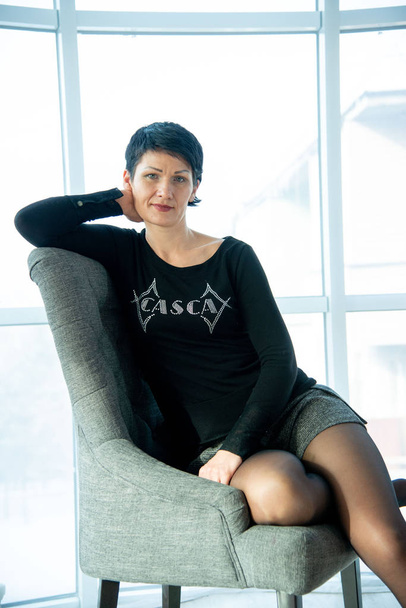portrait brunet woman in black sweater siting in gray soft armchair in studio - Zdjęcie, obraz