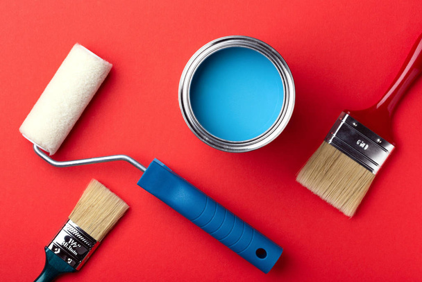 blikje blauwe verf, verfroller en penselen op rood. - Foto, afbeelding