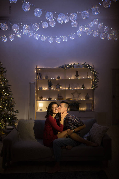 happy, couple in love in the dark, evening on the couch, dark background, christmas tree lights. Christmas evening. New Year. hug and kiss. attraction,romantic evening and date. - Foto, Imagem