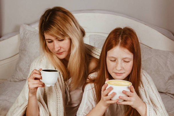 New Year 2020. Mom and ginger-haired daughter are sitting on the bed and drinking cocoa with froth. Covered with a blanket, hug. - Zdjęcie, obraz