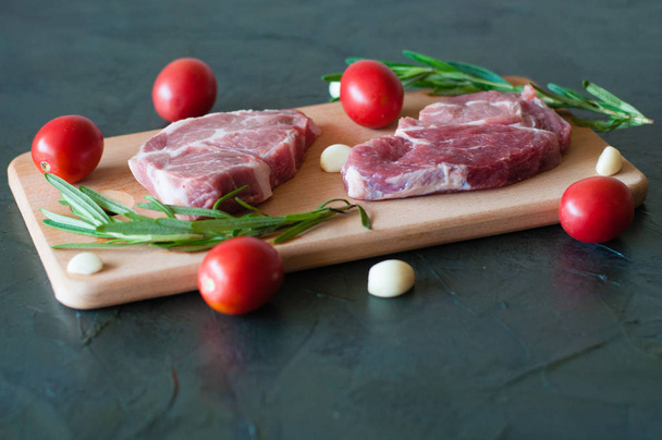 Syrové vepřové steaky s červenými rajčaty, rozmarýnem a česnekem na řezací desce. - Fotografie, Obrázek