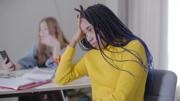 Portrait of irritated African American girl with dreadlocks holding head with hand as blurred Caucasian teen singing and dancing at the table at the background. Students, communication, tiredness. - Materiaali, video