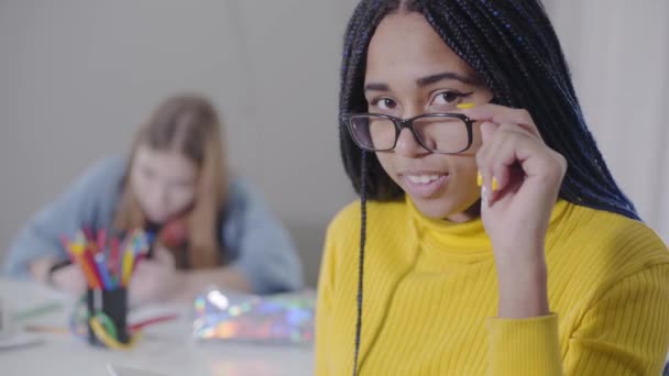 Close-up of beautiful African American woman looking at camera and holding eyeglasses. Cute girl in yellow sweater posing while her Caucasian friend studying at the background. - Video