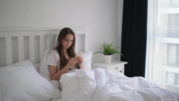 woman is using smartphone lying in bed in morning time - Video