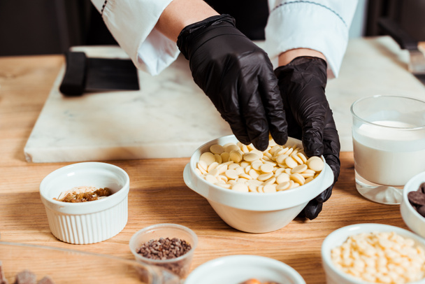 cropped view of chocolatier in black latex gloves taking white chocolate chips  - Foto, Imagen
