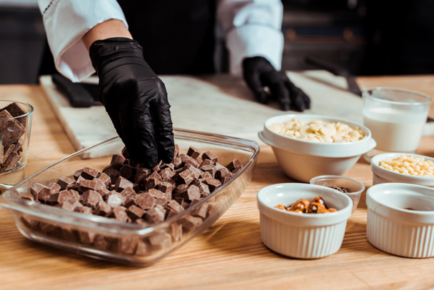 cropped view of chocolatier in black latex gloves taking dark chocolate cubes - Foto, afbeelding