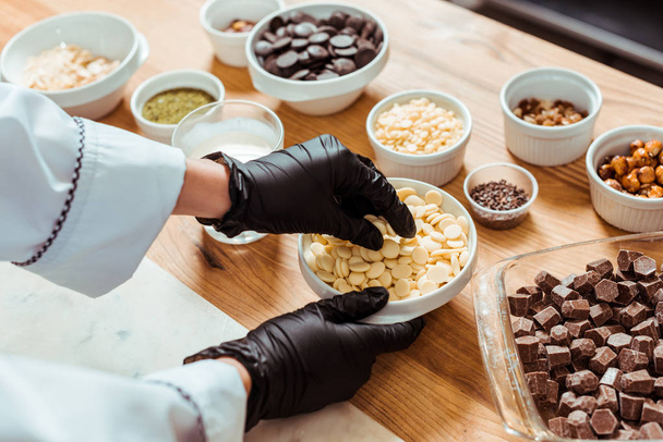 cropped view of chocolatier in black latex gloves taking white chocolate chips near bowls  - Fotoğraf, Görsel
