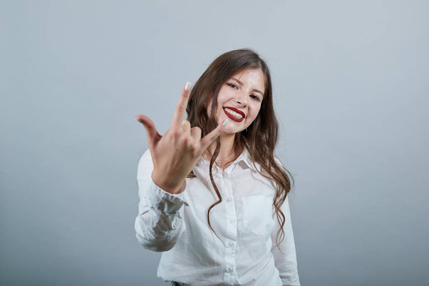 Χαρούμενη όμορφη νεαρή γυναίκα στη μόδα λευκό πουκάμισο κάνει ροκ χειρονομία, χαμογελώντας - Φωτογραφία, εικόνα
