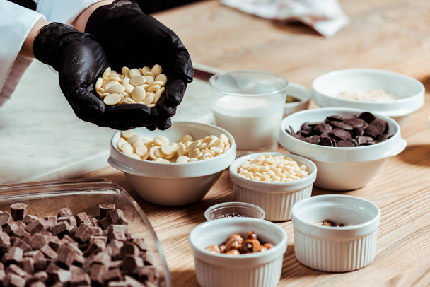 selective focus of chocolatier in black latex gloves holding white chocolate chips - Photo, Image