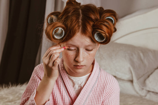 Young beautiful girl does makeup on the bed in the bedroom. Red-haired and caulking with curlers on his head in a bathrobe. - 写真・画像