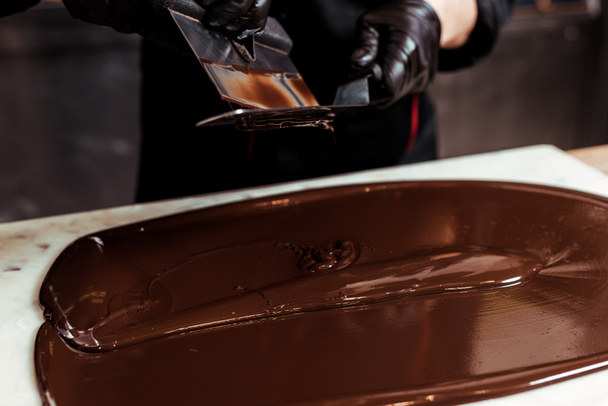 cropped view of chocolatier holding cake scrapers near melted dark chocolate on surface  - Zdjęcie, obraz