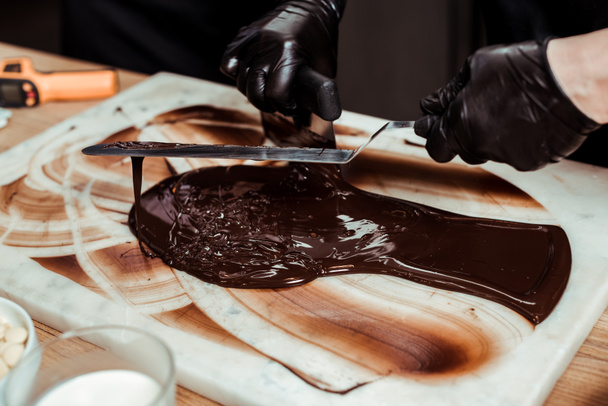 bijgesneden weergave van chocolatier met taart scraper in de buurt gesmolten pure chocolade op het oppervlak  - Foto, afbeelding