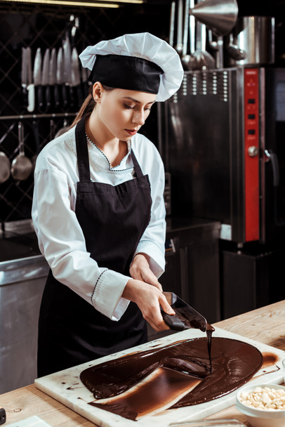 attractive chocolatier holding cake scrapers near melted dark chocolate on marble surface  - Photo, Image