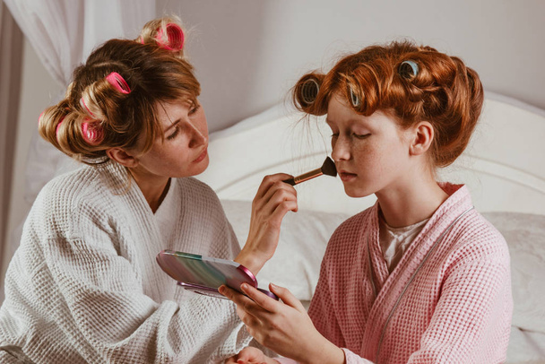 Happy family. Young beautiful mother does makeup of her beautiful daughter on the bed in the bedroom. The red-haired daughter with curlers on her head is laughing. - Photo, Image