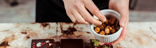 panoramic shot of chocolatier holding bowl with caramelized hazelnuts near chocolate bars  - Photo, Image