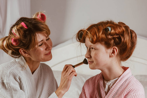 Happy family. Young beautiful mother does makeup of her beautiful daughter on the bed in the bedroom. The red-haired daughter with curlers on her head is laughing. - Φωτογραφία, εικόνα