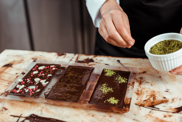 cropped view of chocolatier adding pistachio powder on dark chocolate bar  - Foto, Imagem