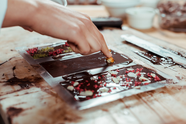vista recortada de chocolatero añadiendo avellanas caramelizadas en barra de chocolate negro
  - Foto, Imagen