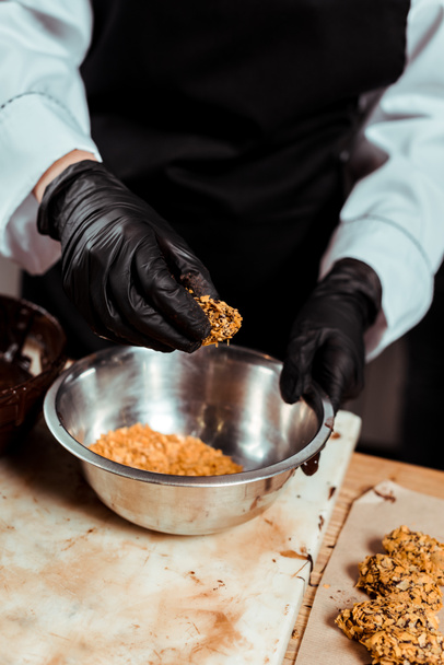 cropped view of chocolatier in black latex gloves holding delicious candy with flakes  - Fotó, kép