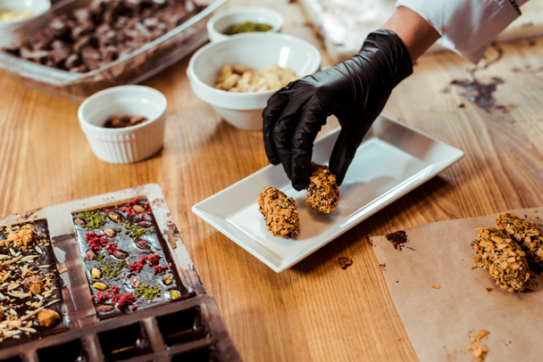 обрізаний вид шоколатьє в чорній латексній рукавичці, що додає смачні цукерки на тарілці
  - Фото, зображення