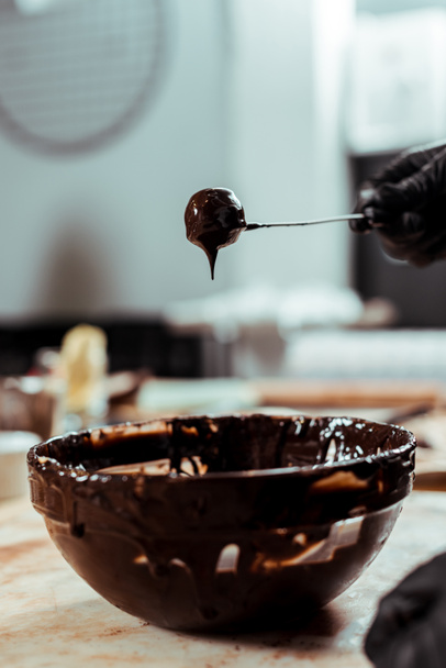vista recortada de chocolatero en guante de látex negro sosteniendo palo con dulces sabrosos cerca de chocolate en un tazón
  - Foto, Imagen