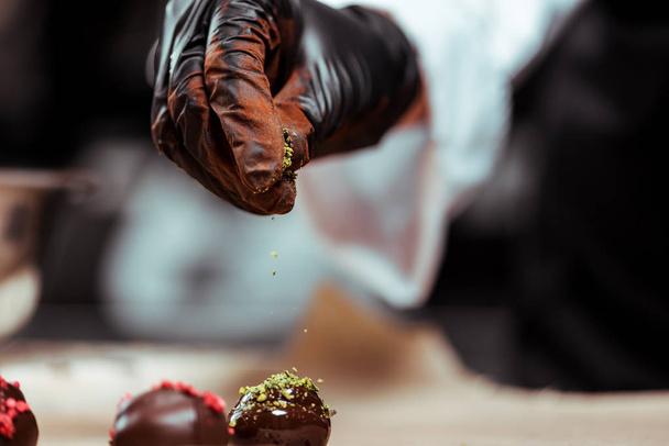 bijgesneden weergave van chocolatier in zwarte latex handschoen toevoegen van groene pistache poeder op vers gemaakte snoepjes  - Foto, afbeelding