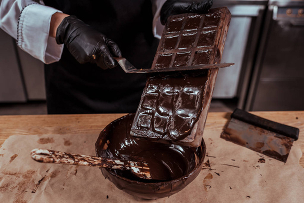 チョコレート金型の近くにケーキスクレーパーを保持するショコラティエのクロップドビューと溶かしたダークチョコレートとボウル  - 写真・画像