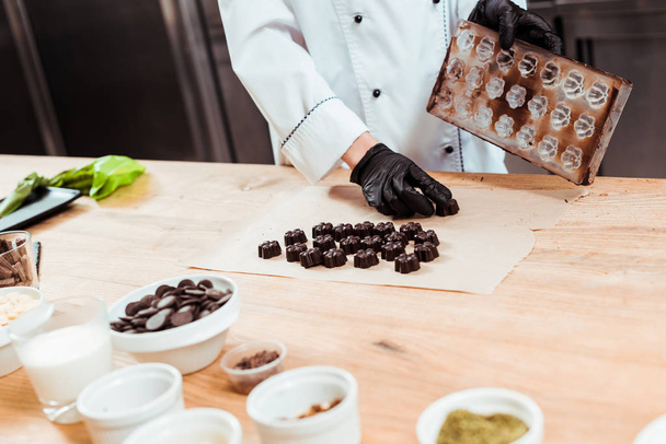 przycięty widok na chocolatier trzymając tacę z lodem w pobliżu przygotowanych cukierków czekoladowych na papierze do pieczenia  - Zdjęcie, obraz