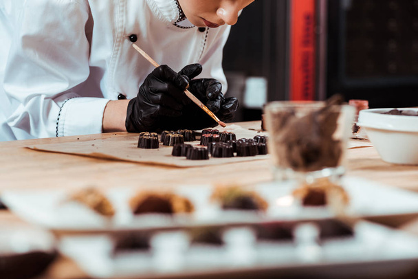 selektiver Fokus des Chocolatiers, der Pinsel mit Goldpuder in der Nähe von vorbereiteten Schokoladenbonbons auf Backpapier hält  - Foto, Bild