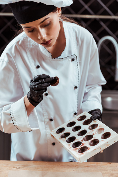 attractive chocolatier looking at tasty candy and holding chocolate molds  - Φωτογραφία, εικόνα