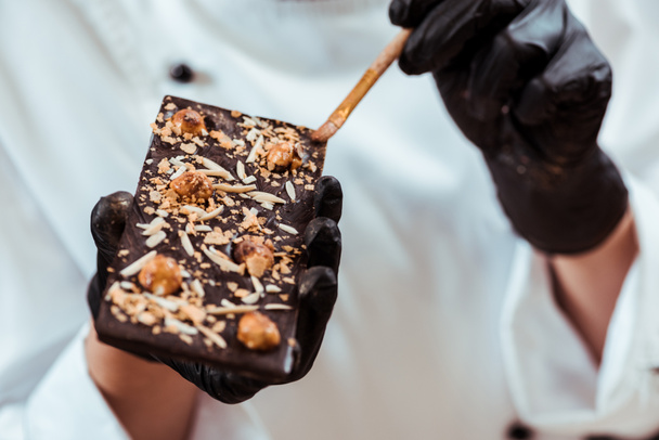 close up of chocolatier holding brush near tasty chocolate bar with hazelnuts  - Foto, afbeelding