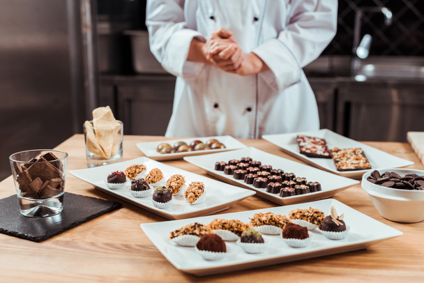 cropped view of chocolatier standing with clenched hands near tasty chocolate candies on plates  - Foto, Imagem