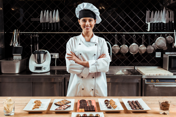 happy chocolatier in chef hat standing with crossed arms near tasty chocolate candies on plates  - Photo, Image