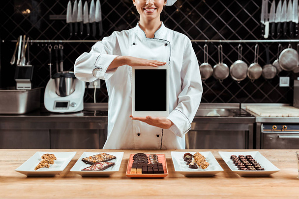 cropped άποψη του χαρούμενα σοκολατένιο στο κρατώντας ψηφιακό δισκίο με λευκή οθόνη κοντά νόστιμα καραμέλες σοκολάτας σε πιάτα  - Φωτογραφία, εικόνα