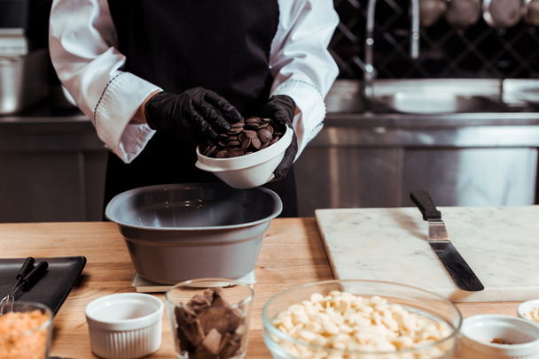 cropped view of chocolatier in latex gloves adding dark chocolate chips into bowl on kitchen scales  - Foto, afbeelding