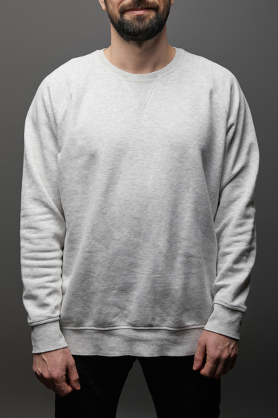 Ausgeschnittene Ansicht eines bärtigen Mannes in leerem grauen Sweatshirt auf schwarzem Hintergrund - Foto, Bild