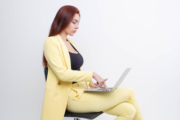 Ένα κοκκινομάλλικο κορίτσι με κίτρινο κοστούμι κάθεται σε μια καρέκλα και δουλεύει σε ένα λάπτοπ. - Φωτογραφία, εικόνα